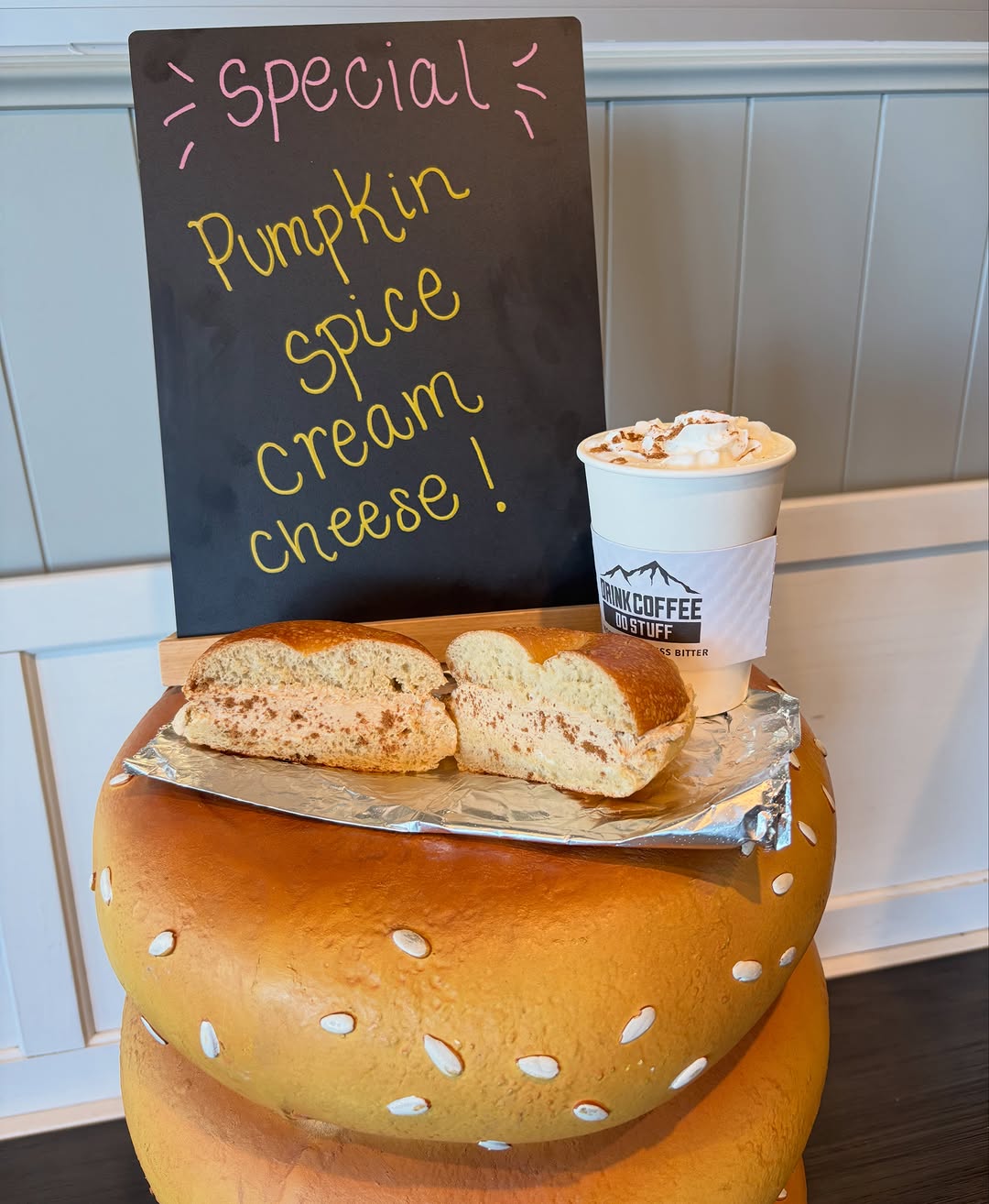A coffee topped with whipped cream next to a bagel with cream cheese, in front of the shop's chalkboard special sign